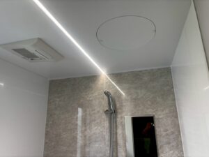 完成した浴室の天井部分。LED照明のラインが壁際に沿って配置され、天井中央には丸型の換気扇が埋め込まれている。壁はグレー系の石目調パネル、天井は白色で明るい印象
