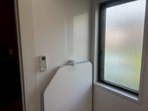 浴室の窓と壁面リモコン。型板ガラスの窓から柔らかな自然光が入り、左側の壁には浴室暖房乾燥機のリモコンが取り付けられている。白色の壁パネルが清潔感を演出